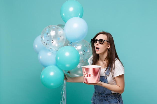 Шокированная женщина в 3D-очках смотрит фильм, держит ведро попкорна, празднуя с красочными воздушными шарами, изолированными на синем бирюзовом фоне. Праздничная вечеринка по случаю дня рождения, эмоции в концепции кино.