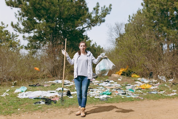 Shocked woman in casual clothes cleaning holding trash bags and rake for garbage collection in littered park