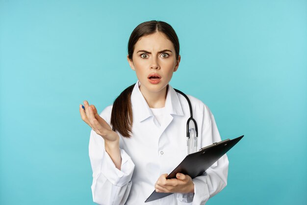 Shocked woman doctor reacting worried and frustrated at camera hear smth strange standing in white coat against torquoise background person