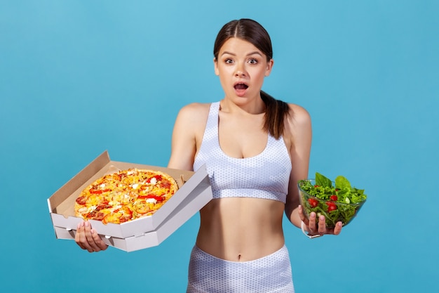 Шокированная женщина, держащая пиццу и миску с салатом из свежих овощей, глядя в камеру большими глазами