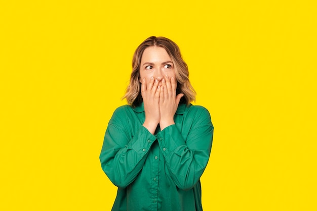 Shocked woman is looking away with her hands covering her mouth wearing a green shirt over yellow wall