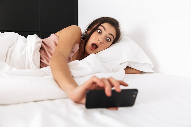 shocked woman looking at mobile phone, while sleeping in bed on white linen