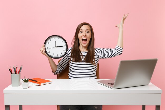 Shocked woman screaming spreading hands holding alarm clock sit, work at office with pc laptop