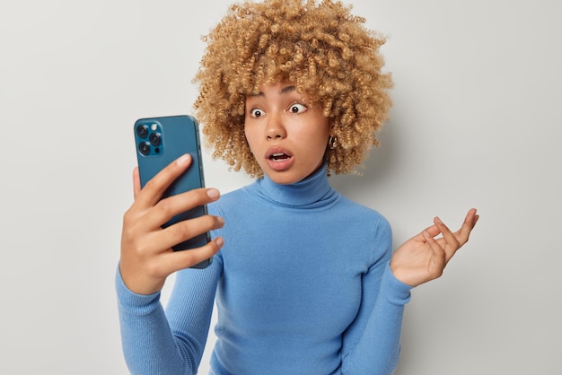 Shocked woman stares at smartphone screen with opened mouth\
keeps hand raised makes video call finds out shocking news wears\
casual blue turtleneck isolated over grey background distant\
communication