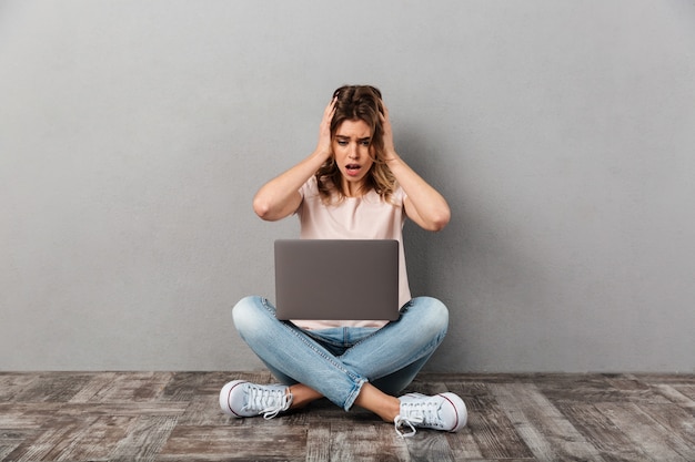 Shocked woman in t-shirt sitting on the floor with laptop computer while holding her head over grey