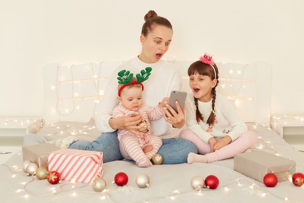 Shocked woman wearing casual clothing sitting on bed with her children, holding cell phone in hands, keeping mouth opened, expressing shock from breaking news.