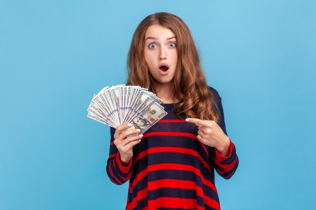 Shocked woman wearing striped casual style sweater pointing at dollars banknotes in her hand having astonished expression big profit Indoor studio shot isolated on blue background