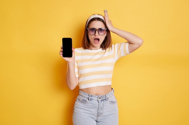 Photo shocked woman wearing striped t shirt and jeans posing isolated over yellow background standing smart phone showing empty display with copy space for promotional test expressing astonishment