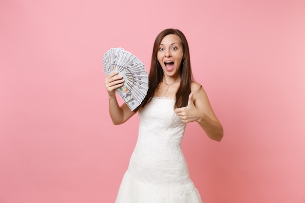 Shocked woman in white dress holding bundle lots of dollars, cash money and showing thumb up