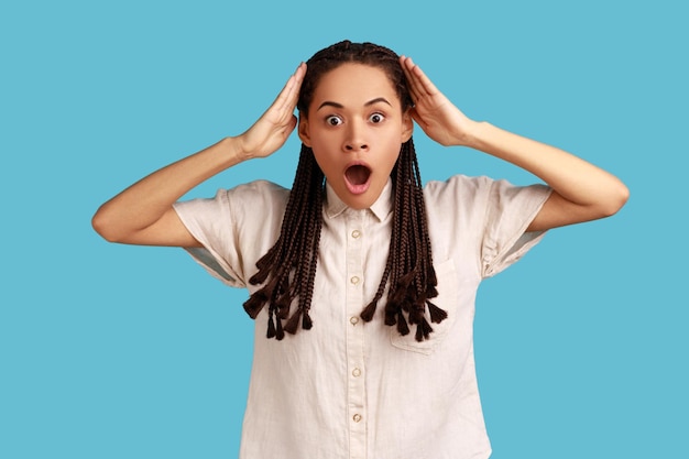 Shocked woman with black dreadlocks reacts on something astonishing raises hands hears surprise news being amazed of something wearing white shirt Indoor studio shot isolated on blue background