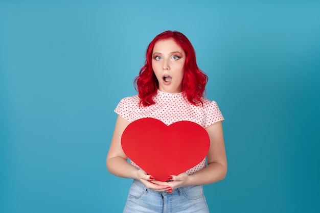 shocked woman with red hair and an open mouth holds a large red paper heart in two hands