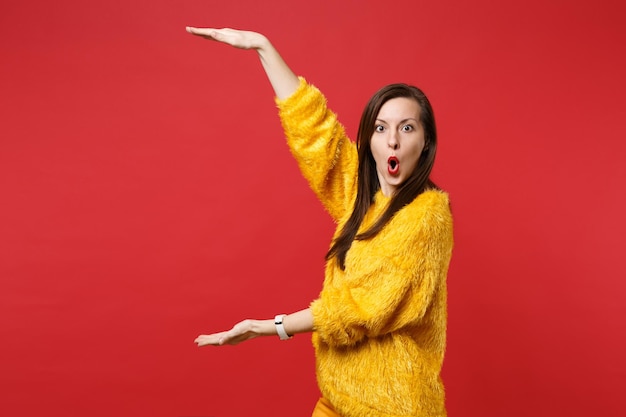노란색 모피 스웨터를 입은 충격을 받은 여성이 입을 벌리고 빨간색 배경에 격리된 수직 작업 공간이 있는 크기를 보여줍니다. 사람들은 진심 어린 감정, 라이프 스타일 개념입니다. 복사 공간을 비웃습니다.