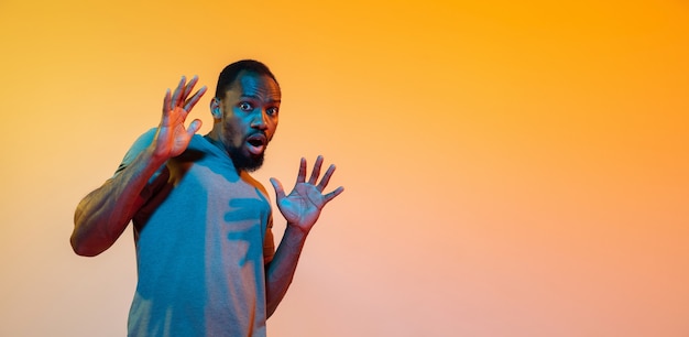 Shocked, wondered. African-american man's modern portrait on gradient orange studio background in neon light. Beautiful afro model. Concept of human emotions, facial expression, sales, ad. Flyer.