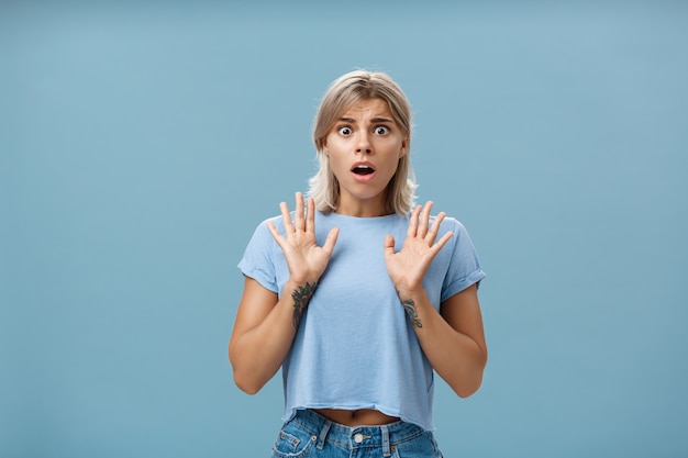 Shocked and worried concerned good-looking feminine woman with blond hair and tattooed arms raising palms gasping and opening mouth expressing empathy and feeling sorry for friend over blue wall