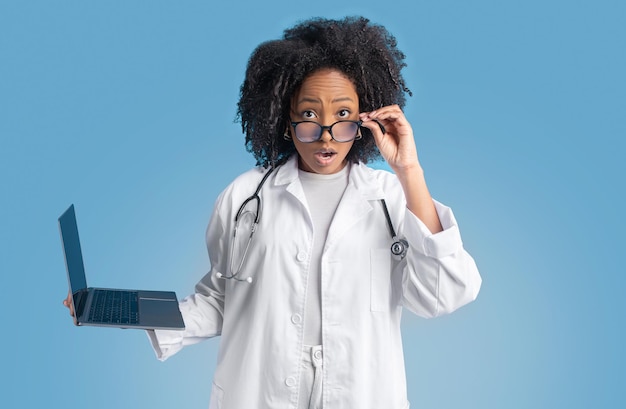 Shocked young african american curly lady doctor therapist in white coat with laptop takes off