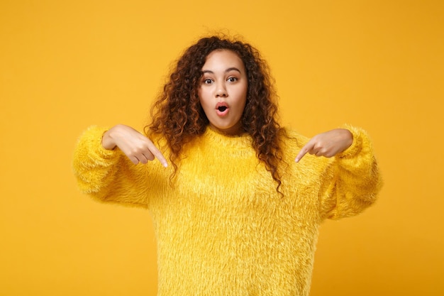 スタジオで黄色のオレンジ色の背景に分離された毛皮のセーターのポーズでショックを受けた若いアフリカ系アメリカ人の女の子。人々の誠実な感情のライフスタイルの概念。コピースペースをモックアップします。人差し指を下に向けます。