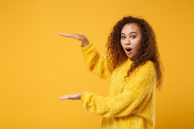 Шокированная молодая афроамериканская девушка в меховом свитере позирует изолированно на желто-оранжевом фоне стены. Концепция образа жизни людей. Скопируйте пространство для копирования. Жесты, демонстрирующие размер с вертикальным рабочим пространством.