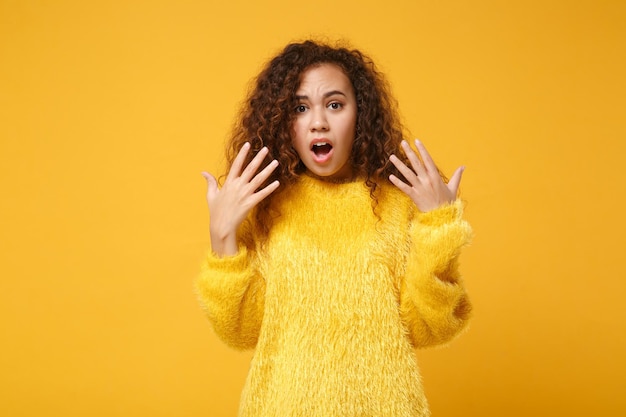 Shocked young african american girl in fur sweater posing isolated on yellow orange wall background, studio portrait. People lifestyle concept. Mock up copy space. Keeping mouth open, spreading hands.