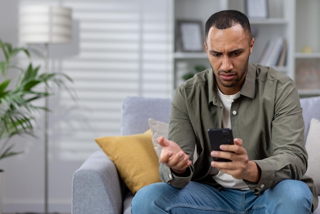 ショックを受けた若いアフリカ系アメリカ人男性が自宅のソファに座り、手に電話を持って心配そうに見ている