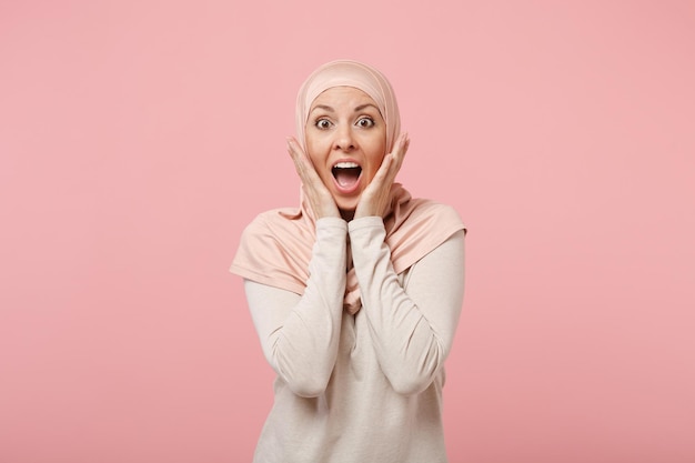 Shocked young arabian muslim woman in hijab light clothes posing isolated on pink background. People religious Islam lifestyle concept. Mock up copy space. Keeping mouth open, putting hands on cheeks.