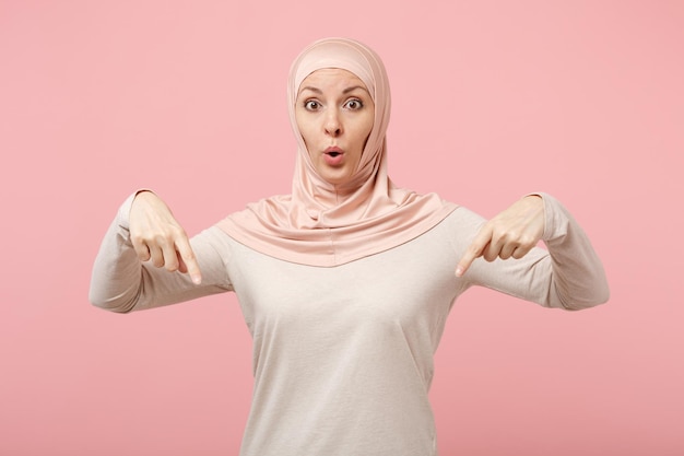 Shocked young arabian muslim woman in hijab light clothes posing isolated on pink wall background in studio. People religious Islam lifestyle concept. Mock up copy space. Pointing index fingers down.