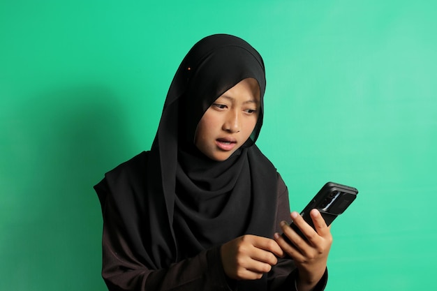 Shocked young Asian girl holding smartphone isolated on green background