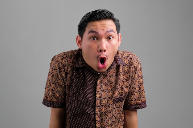 Шокированный молодой азиат в рубашке из батика смотрит в камеру с открытым ртом на сером фоне