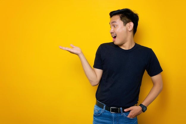 Shocked young Asian man in casual tshirt showing copy space on palm isolated on yellow background Great offer concept