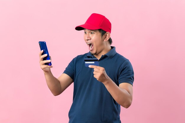 Foto giovane uomo asiatico scioccato in maglietta casual con un cappello che tiene il telefono cellulare e la carta di credito
