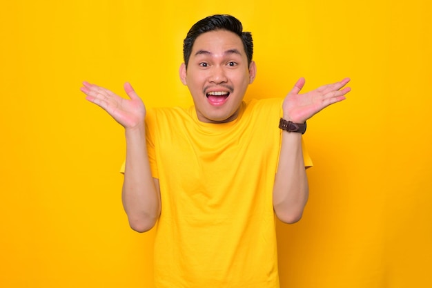 Shocked young Asian man in casual tshirt with open mouth and raising his palms up isolated on yellow background People lifestyle concept