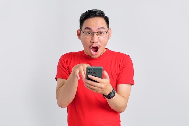 Shocked young Asian man holding mobile phone and looking at camera with opened mouth isolated over white background