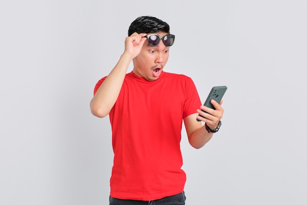 Shocked young Asian man looking at mobile phone screen reacting to online news with opened mouth isolated over white background