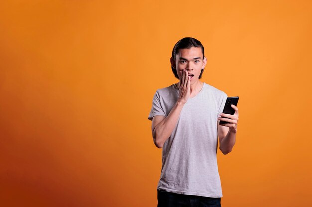 Shocked young asian man looking at smartphone, reading news online, medium shot. teenager standing with open mouth and hand on face, holding mobile phone, hearing gossips