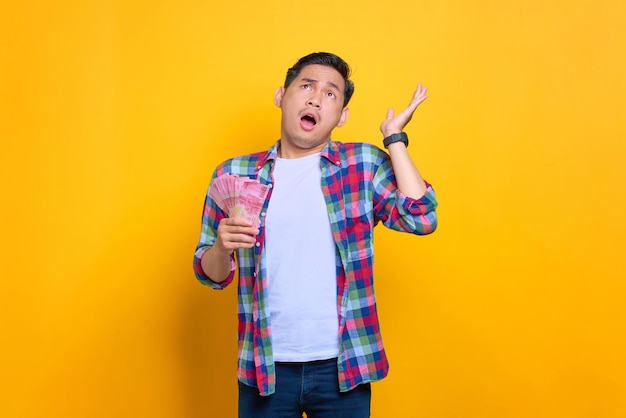 Shocked young Asian man in plaid shirt holding money banknotes and looking up isolated on yellow background