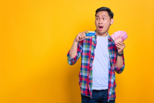 Shocked young Asian man in plaid shirt showing money banknotes and credit card looking away at copy space isolated on yellow background