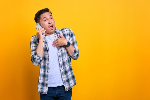 Shocked young Asian man in plaid shirt talking on mobile phone looking aside at copy space isolated on yellow background