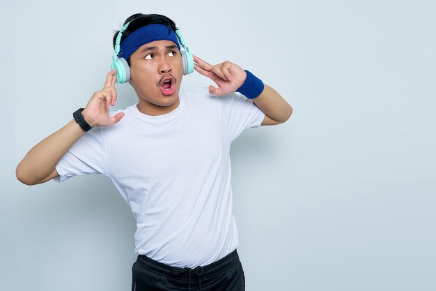 Shocked young asian man sporty fitness trainer instructor in blue headband and white tshirt Listen to music with headphones isolated over white background