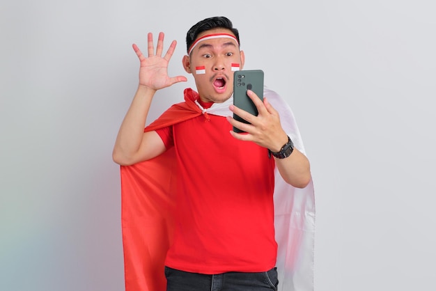 흰색 배경 인도네시아 독립 기념일 개념에 격리된 휴대 전화를 사용하여 충격을 받은 젊은 아시아 남자