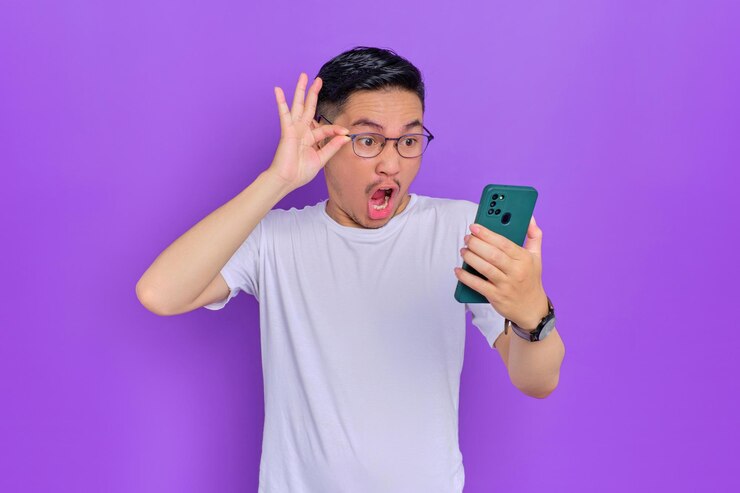 白い t シャツにスマートフォンを使用し、紫色の背景に分離されたメガネを脱いでショックを受けた若いアジア人男性