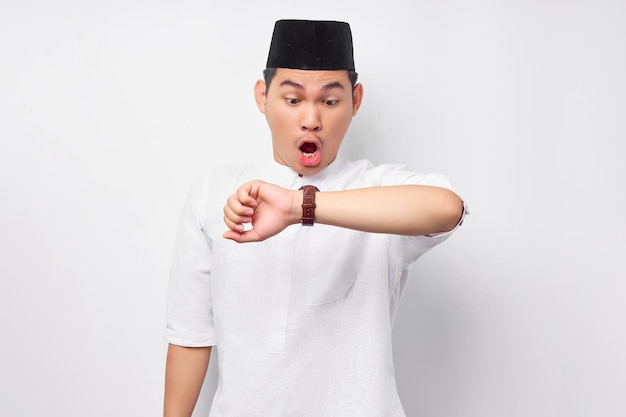 아랍 옷을 입은 젊은 아시아계 무슬림 남성이 팔을 들고 흰 배경에 격리된 손목시계를 보고 있다.
