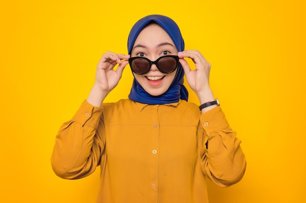 Shocked young asian muslim woman dressed in orange shirt\
looking at camera with hand touching his glasses interested to big\
promo isolated on yellow background