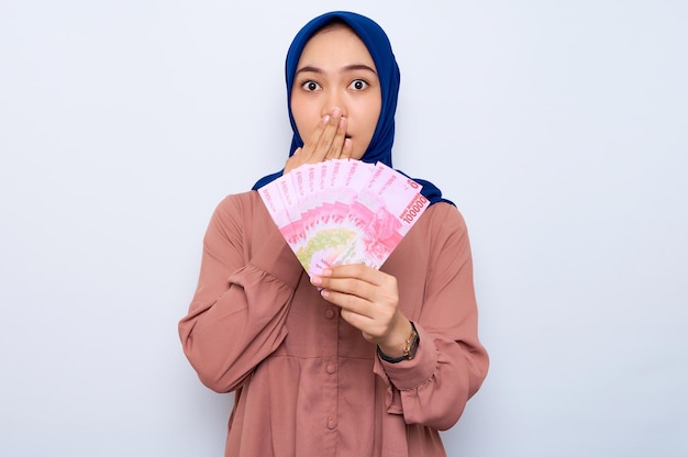 Шокированная молодая азиатка-мусульманка в розовой рубашке с денежными банкнотами, изолированными на белом фоне Концепция религиозного образа жизни людей