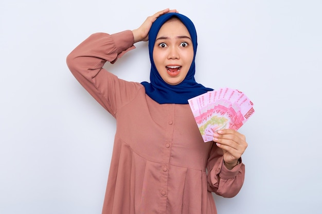 Shocked young Asian muslim woman in pink shirt holding money banknotes isolated over white background People religious lifestyle concept