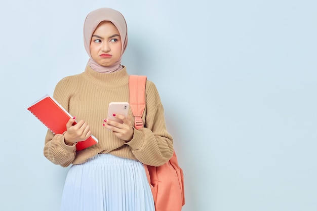 Shocked young asian muslim woman student in brown sweater with\
backpack holding book and mobile phone isolated on white background\
back to school concept