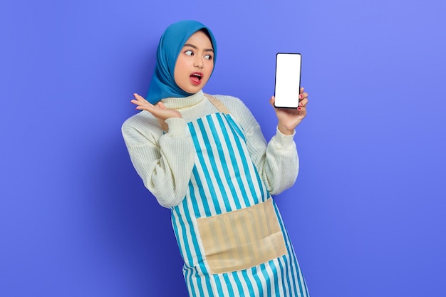 히잡과 앞치마를 입은 젊은 아시아 이슬람 여성이 보라색 배경에 격리된 손으로 빈 화면 휴대전화를 들고 충격을 받았습니다. 사람들이 주부 이슬람 라이프 스타일 개념