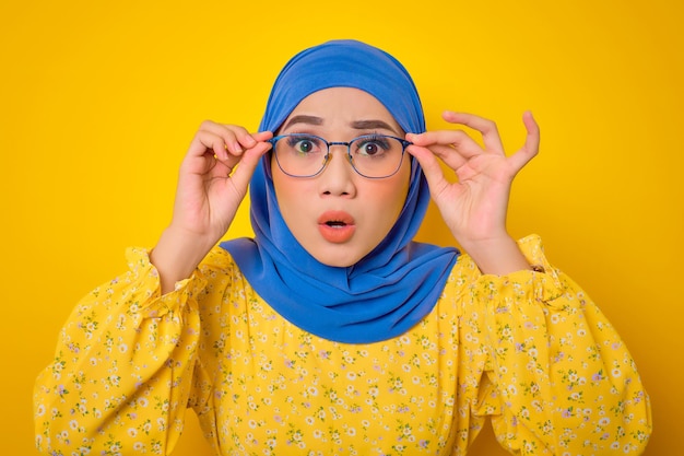 Shocked young Asian woman in casual dress touching his glasses with mouth opened emotionally reacting to news isolated on yellow background