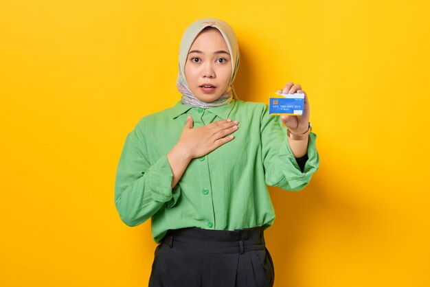 Shocked young Asian woman in a green shirt showing credit card on yellow background