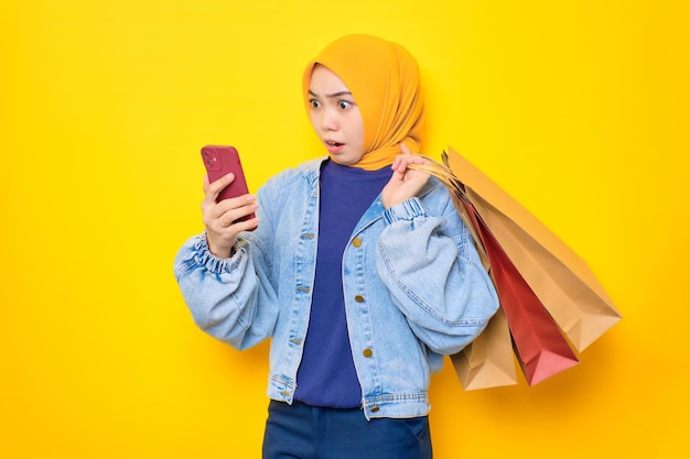 노란색 배경에 격리된 쇼핑백을 들고 온라인 쇼핑을 위해 휴대전화를 사용하는 청바지 재킷을 입은 젊은 아시아 여성