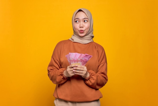 Shocked young Asian woman in orange shirt holding money banknotes and looking up isolated over yellow background