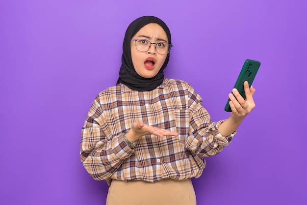 Shocked young Asian woman in plaid shirt holding a mobile phone getting great news looking at camera isolated on purple background